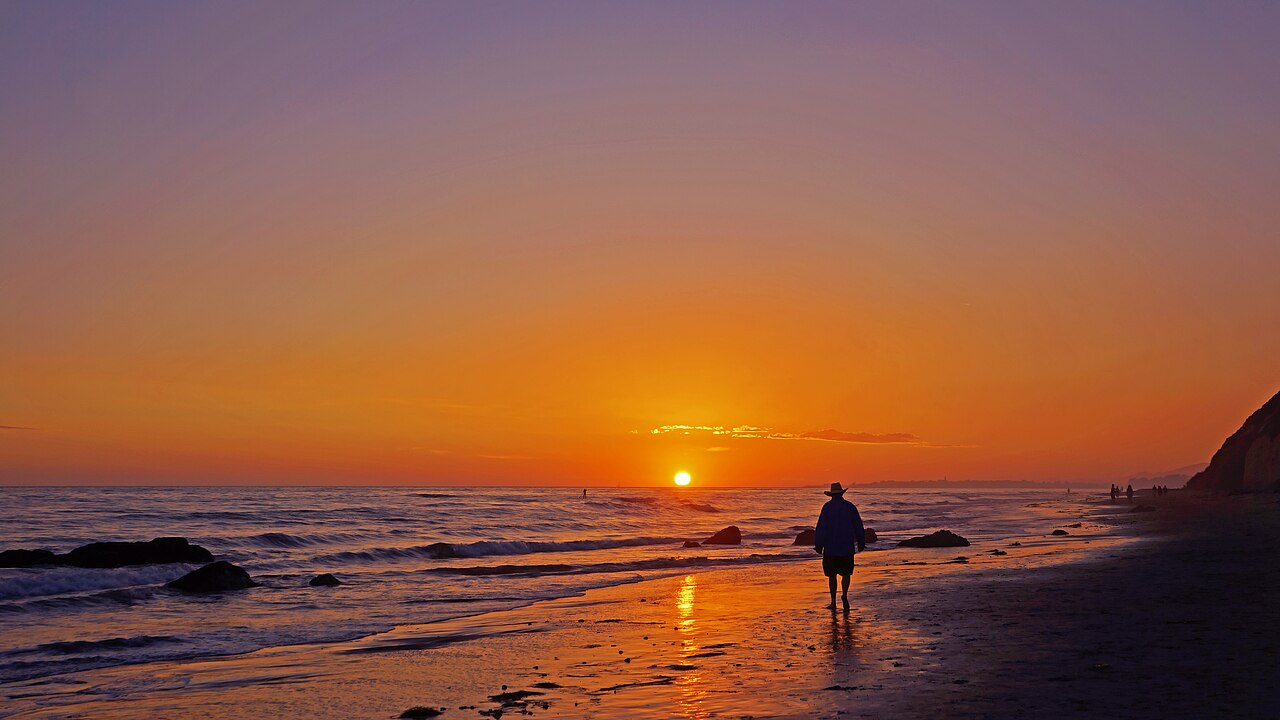 A working cowboy on horseback silhouetted against a fiery orange Texas sunset, hat tipped low, lasso coiled at the saddle.
